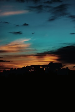 The image displays a picturesque sunset featuring a gradient sky with hues of blue transitioning into warm oranges and yellows near the horizon. Dark clouds and silhouetted shapes of buildings or trees are present at the bottom, creating a striking contrast against the illuminated sky.