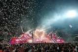A large crowd gathers in front of a brightly lit stage with colorful lights, lasers, and smoke effects filling the air. Confetti falls from above, creating a festive and energetic atmosphere. People are holding up their phones and enjoying the event.