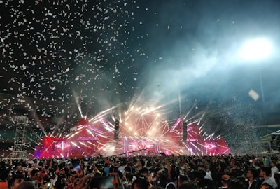 A large crowd gathers in front of a brightly lit stage with colorful lights, lasers, and smoke effects filling the air. Confetti falls from above, creating a festive and energetic atmosphere. People are holding up their phones and enjoying the event.