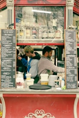 Two people are working in a small outdoor kiosk or food stand. The kiosk has a menu on either side listing various items and prices. Several bottles of beverages are visible through a glass panel above the workers. The individuals are focused on preparing food or drinks.