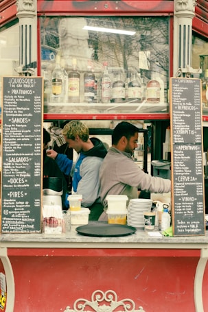 Two people are working in a small outdoor kiosk or food stand. The kiosk has a menu on either side listing various items and prices. Several bottles of beverages are visible through a glass panel above the workers. The individuals are focused on preparing food or drinks.