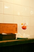 A wine glass filled with red liquid and fruit slices sits on a counter next to a wooden box and a menu standing upright. There is also a plate with crumpled napkins next to these items. The background consists of a wall with white tiles.