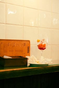 A wine glass filled with red liquid and fruit slices sits on a counter next to a wooden box and a menu standing upright. There is also a plate with crumpled napkins next to these items. The background consists of a wall with white tiles.