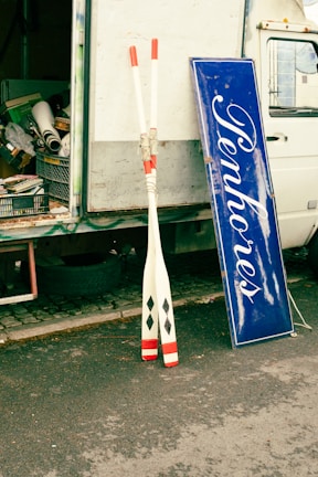 A pair of oars with red and black markings is leaning against the open back of a truck. Nearby, a blue sign with white cursive text is propped up, and various items and equipment can be seen inside the truck.