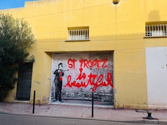 A large mural on a yellow wall features a black and white figure holding a paint roller with splashes of red paint. The words 'St Tropez is Beautiful' are boldly written in red graffiti style. The setting appears to be a quiet street with some greenery on the left side and a few barred windows above.
