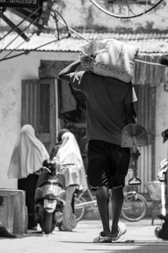 A person carrying a large cement bag on their shoulder walks down a street, holding a shovel. In the background, two individuals wearing traditional long garments are standing next to a parked motorcycle. The scene is set in an outdoor urban area with various structures and wires visible.