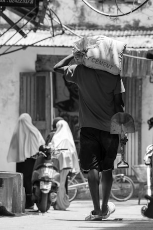 A person carrying a large cement bag on their shoulder walks down a street, holding a shovel. In the background, two individuals wearing traditional long garments are standing next to a parked motorcycle. The scene is set in an outdoor urban area with various structures and wires visible.