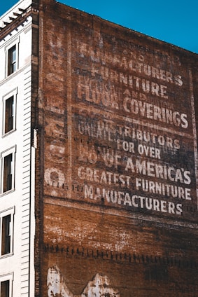 A vintage advertisement painted on the side of a brick building, featuring words like 'manufacturers', 'furniture', and 'floor coverings'. The text is slightly faded, indicating weathering over time. The adjacent part of the building is white and features windows, providing a contrast to the brown brick wall.