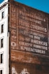A vintage advertisement painted on the side of a brick building, featuring words like 'manufacturers', 'furniture', and 'floor coverings'. The text is slightly faded, indicating weathering over time. The adjacent part of the building is white and features windows, providing a contrast to the brown brick wall.