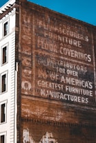 A vintage advertisement painted on the side of a brick building, featuring words like 'manufacturers', 'furniture', and 'floor coverings'. The text is slightly faded, indicating weathering over time. The adjacent part of the building is white and features windows, providing a contrast to the brown brick wall.