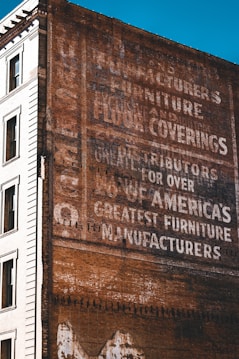 A vintage advertisement painted on the side of a brick building, featuring words like 'manufacturers', 'furniture', and 'floor coverings'. The text is slightly faded, indicating weathering over time. The adjacent part of the building is white and features windows, providing a contrast to the brown brick wall.