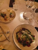 A dining table set with plates featuring gourmet dishes, including one with sliced meat topped with greens and cheese shavings, and another with assorted small servings and a dollop of butter. Two wine glasses are partially visible on the table, along with a lit candle in a glass holder and a glass of water.