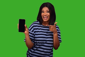 A smiling person wearing a striped shirt enthusiastically points at a smartphone they are holding against a bright green background.