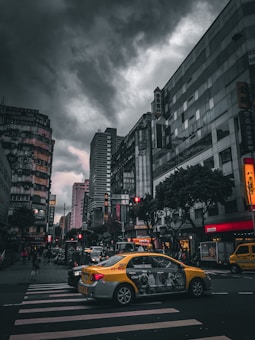 A bustling urban street scene featuring tall buildings on either side. A yellow taxi is prominently displayed in the foreground as it drives through a crosswalk. Pedestrians and other vehicles are visible on the street, and the sky is overcast with dark, dramatic clouds looming overhead. The use of muted colors and shadows creates a moody, atmospheric effect.