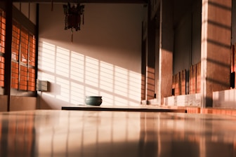 Warm sunlight casts long shadows through a window with lattice patterns, illuminating a minimalist interior space. A small green ceramic bowl sits on a smooth, reflective surface, adding a focal point to the serene setting. The walls and window frames are made of wood, complemented by a traditional hanging ornament near the ceiling.