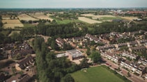 Aerial view of a suburban neighborhood surrounded by lush greenery and farmland. Rows of houses with gable roofs are organized along curving streets. Open fields and patches of forest can be seen beyond the residential area.