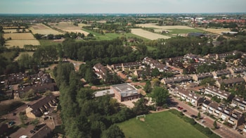 Aerial view of a suburban neighborhood surrounded by lush greenery and farmland. Rows of houses with gable roofs are organized along curving streets. Open fields and patches of forest can be seen beyond the residential area.