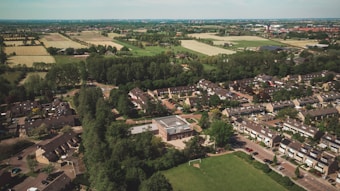 Aerial view of a suburban neighborhood surrounded by lush greenery and farmland. Rows of houses with gable roofs are organized along curving streets. Open fields and patches of forest can be seen beyond the residential area.