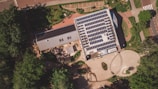 An aerial view of a building with a large array of solar panels on its roof. The structure is surrounded by greenery, pathways, and some smaller structures or landscaped areas. The environment appears to be a mix of natural and man-made elements, suggesting a focus on sustainability.