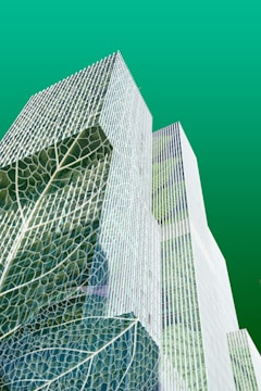 Tall, modern skyscrapers are creatively overlaid with large leaf textures, suggesting an integration of nature with urban architecture against a smooth green background.