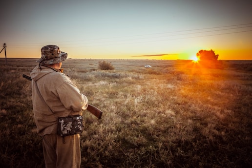 A person wearing a camouflage hat and jacket stands in an open field, holding a rifle. The sun is setting on the horizon, casting a warm glow across the landscape. A small white vehicle is visible in the distance near a tree. The sky is clear with few clouds.
