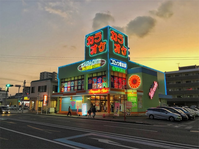 A neon-lit karaoke building with vibrant signs is seen during sunset. The signs display Japanese characters and words like 'Karaoke' and 'Open 24h'. The structure is surrounded by parked cars and people walking by, with a background of other buildings and a clear evening sky.