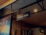 A well-lit ordering area in a beverage shop with a prominent 'ORDER HERE' sign hanging from the ceiling. The background includes a single menu board displaying various drinks and their prices, along with some metallic containers and a plant visible in the upper left. The ambiance features a combination of industrial and modern design elements, with a wire mesh and warm lighting.