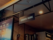 A well-lit ordering area in a beverage shop with a prominent 'ORDER HERE' sign hanging from the ceiling. The background includes a single menu board displaying various drinks and their prices, along with some metallic containers and a plant visible in the upper left. The ambiance features a combination of industrial and modern design elements, with a wire mesh and warm lighting.