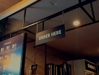 A well-lit ordering area in a beverage shop with a prominent 'ORDER HERE' sign hanging from the ceiling. The background includes a single menu board displaying various drinks and their prices, along with some metallic containers and a plant visible in the upper left. The ambiance features a combination of industrial and modern design elements, with a wire mesh and warm lighting.