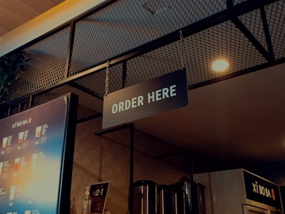 A well-lit ordering area in a beverage shop with a prominent 'ORDER HERE' sign hanging from the ceiling. The background includes a single menu board displaying various drinks and their prices, along with some metallic containers and a plant visible in the upper left. The ambiance features a combination of industrial and modern design elements, with a wire mesh and warm lighting.