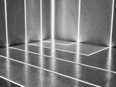 Modern minimalist interior featuring a tiled floor and walls with thin illuminated lines accentuating the geometric patterns. The lighting creates a reflective surface on the tiles, enhancing the clean and sleek design.