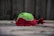 A worn-out tennis ball lies on a surface with its outer layer torn, revealing green and red fuzz. The background appears blurred with a fence-like structure.