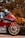 A red sport motorcycle with gold wheels is parked on a road next to a concrete barrier. The background features autumn-colored trees, creating a warm, rustic setting. The motorcycle has a sleek design with a noticeable Honda logo.
