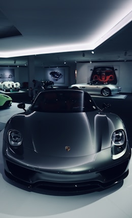 A sleek, modern sports car in a dimly lit showroom, reflecting ambient lights. The glossy finish enhances its aerodynamic design. Other luxury cars are visible in the background, adding to the scene's sophistication.