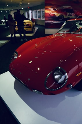 A vintage red sports car is prominently displayed on a white platform in an indoor showroom. In the background, another yellow sports car is visible, with people standing nearby observing the vehicles. The walls are adorned with motor racing-themed artwork, adding to the automotive ambiance.