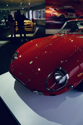 A vintage red sports car is prominently displayed on a white platform in an indoor showroom. In the background, another yellow sports car is visible, with people standing nearby observing the vehicles. The walls are adorned with motor racing-themed artwork, adding to the automotive ambiance.
