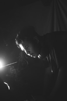 A calm, focused person sitting thoughtfully in a dimly lit room with a dark background and soft light highlighting their face.