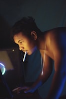 A person with short hair is illuminated by soft lighting while leaning forward towards a computer. They have a cigarette in their mouth and are shirtless, wearing a chain necklace. The lighting creates a dramatic effect with contrasts of blue and warm tones.