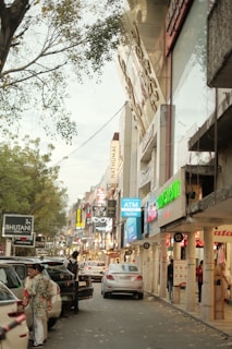 A bustling urban street lined with various shops displaying bright, colorful signs. Cars are parked along the roadside while pedestrians are seen walking and engaging with storefronts. Overhead, tree branches add a natural touch to the busy city scene.