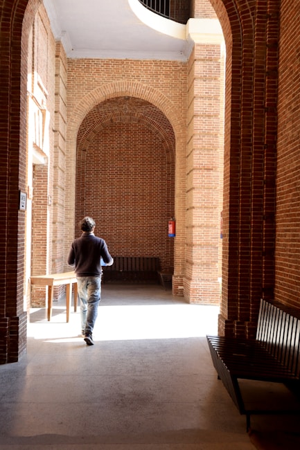 A person walks through a hallway made of red brick arches. The corridor is illuminated by natural light creating a contrast of shadows on the stone floor. Benches are placed along the walls, and a wooden table is visible on the left. The atmosphere is serene and contemplative.