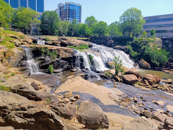 A series of cascading waterfalls flows over rocky terrain surrounded by lush greenery. In the background, modern buildings rise above the treetops, merging nature with urban development.