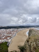 A coastal city with many buildings featuring red-tiled roofs. A wide, sandy beach runs along the coastline, bordering the city. Cliffs are present in the foreground with some sparse vegetation and overcast sky with thick clouds covering the area. The sea is visible on the right with gentle waves washing onto the beach.