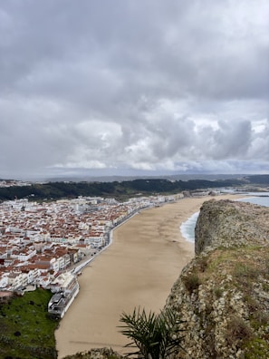 A coastal city with many buildings featuring red-tiled roofs. A wide, sandy beach runs along the coastline, bordering the city. Cliffs are present in the foreground with some sparse vegetation and overcast sky with thick clouds covering the area. The sea is visible on the right with gentle waves washing onto the beach.