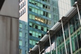 A modern architectural scene featuring glass skyscrapers reflecting nearby buildings. The structures display a mix of geometric shapes and lines, with windows casting a greenish tint. The architectural design is sleek and contemporary, with metal and glass surfaces contributing to an urban aesthetic.