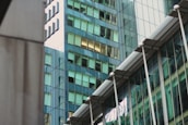 A modern architectural scene featuring glass skyscrapers reflecting nearby buildings. The structures display a mix of geometric shapes and lines, with windows casting a greenish tint. The architectural design is sleek and contemporary, with metal and glass surfaces contributing to an urban aesthetic.