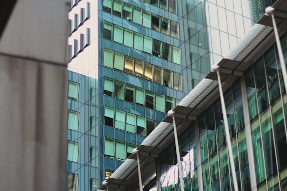 A modern architectural scene featuring glass skyscrapers reflecting nearby buildings. The structures display a mix of geometric shapes and lines, with windows casting a greenish tint. The architectural design is sleek and contemporary, with metal and glass surfaces contributing to an urban aesthetic.