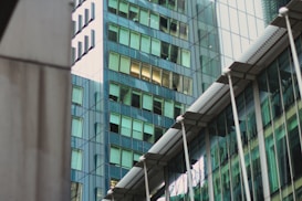 A modern architectural scene featuring glass skyscrapers reflecting nearby buildings. The structures display a mix of geometric shapes and lines, with windows casting a greenish tint. The architectural design is sleek and contemporary, with metal and glass surfaces contributing to an urban aesthetic.