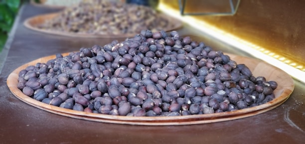 A wooden tray filled with a large pile of dark brown and black coffee beans placed on a brown surface, with more blurred trays of the same in the background.