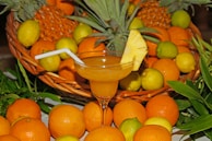 A vibrant display of fresh citrus fruits, including oranges, lemons, and limes, surrounds a basket. A tropical drink garnished with a pineapple slice and a straw is prominently featured in a cocktail glass in the foreground.