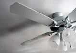 A close-up view of a ceiling fan with four blades and attached light fixtures. The fan and light fixtures are white, with the light fixtures having a floral, frosted glass design. Two pull chains hang from the light fixture. The fan is mounted on a smooth ceiling, casting subtle shadows.